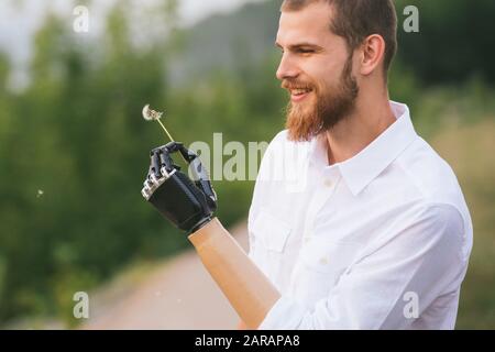 Mann hält Löwenzahn in einer bionischen Hand und betrachtet es. Seitenansicht. Stockfoto
