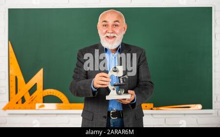 Glücklicher Oberlehrer verwendet Mikroskop. Chemie im Schullabor. Wissenschaftliche Experimente mit Mikroskop im Labor. Laborgeräte für Biologieschulen. Mann im Klassenzimmer mit Mathematikwerkzeugen an der Tafel. Stockfoto