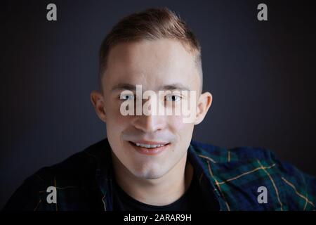 Attraktives Gesicht eines jungen Mannes kaukasischer Ethnie in Nahaufnahme auf schwarzem Hintergrund. Freundliches Lächeln eines weißen Mannes vor einem dunklen Zimmer. Charme Stockfoto