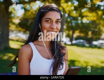 Porträt einer lächelnden jungen Frau mit Ohrhörern in den Ohren Blick auf die Kamera im Park - Fröhliche junge Frau, die im Freien Musik hört Stockfoto