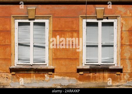 Zwei geschlossene verfallene Fenster mit weißen Holzrahmen und kaputten Kunststoff-Fensterjalousien, die an der Wand des alten Vorstadt-Familienhauses angebracht sind Stockfoto