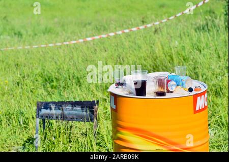 Zerbrochenes und gebrauchenes Einmal-Geschirr aus Kunststoff auf einem Metall-Brennstoff-Fass vor dem Hintergrund von grünem Gras an einem klaren Sommertag Russland, Kursker Region, Zh Stockfoto