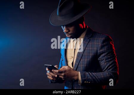 Porträt eines selbstbewussten jungen afrikaners in formaler Kleidung, Festhalten des Mobiltelefons, Blick auf den Bildschirm, stehen vor schwarzem Hintergrund. Erfolgreich Stockfoto