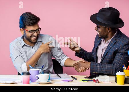 Glückliche afroamerikanische und indische Geschäftspartner sitzen am Schreibtisch im Büro und schütteln die Hände, fröhlich lachen, genießen viel und Erfolg. Stockfoto