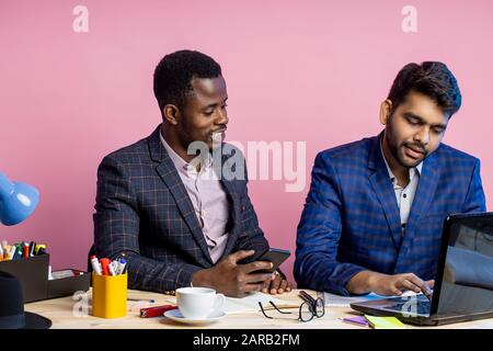 Schuss von zwei Geschäftsleuten, die Anzüge im Büro am Schreibtisch mit Laptop und Handy tragen. Indianischer Mann, der Ideen mit einem afroamerikanischen Kollegen teilt, Stockfoto