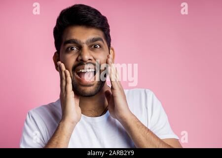Verblüffte, unrasierte indische Männer, schockiert von niedrigen Verkaufspreisen, händeringend auf Wangen, überraschenderweise Mundöffnung, isoliert über rosafarbenem Bac Stockfoto