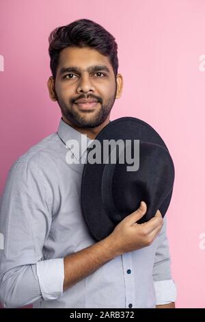 Vertikaler Schuss des selbstbewussten, ruhigen jungen, unrasierten Indianers in grauem Hemd, mit modischem Haarschnitt, mit schwarzem Hut in der Hand und Blick auf den Kamer Stockfoto