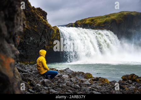 Tourist in einer gelben Jacke, entspannend am Wasserfall Godafoss in Island Stockfoto