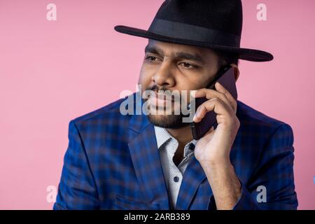 Besorgter junger indischer bärtiger Geschäftsmann, der auf dem Handy spricht. Nahaufnahme des Mannes in formeller Kleidung, schwarzer Hut, mit ernsthaftem, konzentriertem Aussehen während ha Stockfoto