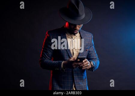 Porträt eines selbstbewussten jungen afrikaners in formaler Kleidung, Festhalten des Mobiltelefons, Blick auf den Bildschirm, stehen vor schwarzem Hintergrund. Erfolgreich Stockfoto
