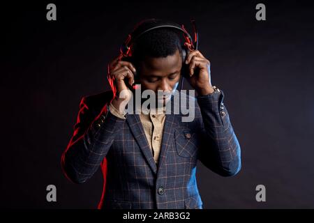 Nahaufnahme des jungen, unshaven afroamerikanischen Mannes, der Musik in Kopfhörern hört, nach unten schaut und im Dunkeln posiert. Porträt eines eleganten DJ mit Headset, Stockfoto