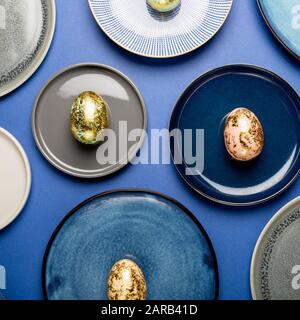 Andere keramische Platten mit goldenen Gesprenkelte Ostereier von Pastellfarben auf blauem Hintergrund. Frohe Ostern Karte Konzept, minimalistisches Design, Ansicht von oben Stockfoto