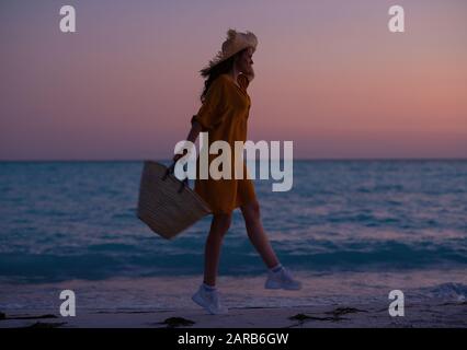 Silhouette der modernen Frau mit langen brünetten Haaren mit Strandtasche, die bei Sonnenuntergang an der Küste springt. Stockfoto