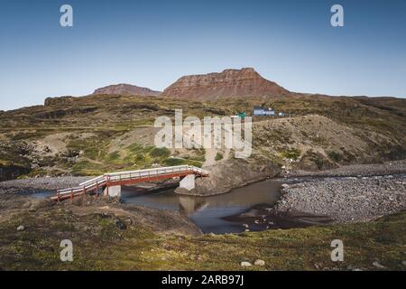 Wanderpfad auf der arktischen Disko-Insel in Grönland. Flussbett mit Brücke und bunten Häusern im Hintergrund. Tafelberge an einem sonnigen Tag im Sommer. Stockfoto