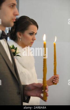 Brautpaar steht bei der Hochzeitsfeier. Fröhliches stilvolles Hochzeitspaar, das während der heiligen Ehe Kerzen mit Licht unter goldenen Kronen hält Stockfoto