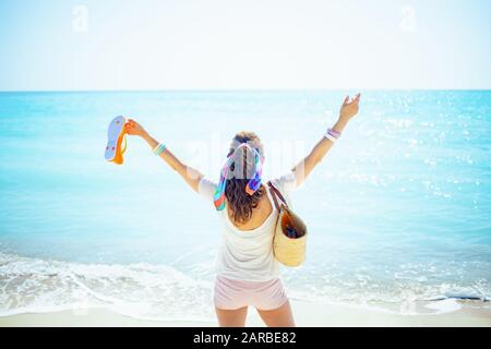 Von der trendigen 40-jährigen Frau in weißem T-Shirt mit Strandabschnitt und orangefarbenen Flip-Flops mit erhobenen Armen am Meeresufer gesehen. Stockfoto