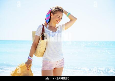 Lächelnde, moderne Frau in weißem T-Shirt mit Strandabdeckung und großem Hut am Strand. Stockfoto