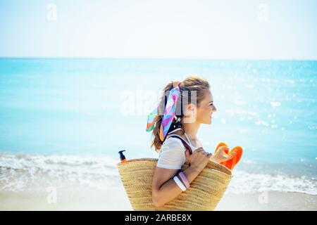 Fröhliche junge Frau in weißem T-Shirt mit Strandabschnitt und orangefarbenen Flip-Flops, die an der Küste spazieren gehen. Stockfoto