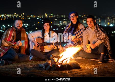 Eine Gruppe von Leuten, die nachts in der Nähe von Lagerfeuer sitzen und Gitarre spielen, Lieder singen und glücklich miteinander sprechen. Stockfoto