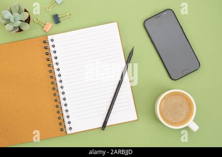 Leeres Notizbuch mit leerer Seite, Kaffeetasse und Hand mit Bleistift. Tischplatte, Arbeitsbereich auf grünem Hintergrund. Kreatives flaches Lay. Stockfoto