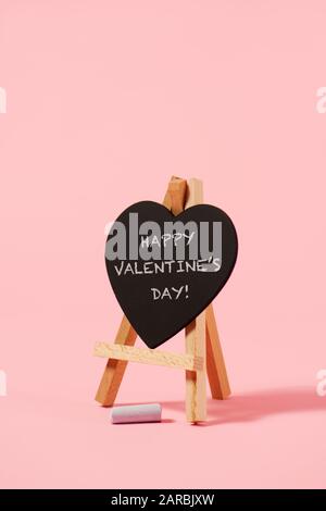 Ein herzförmiges schwarzes Kelch, das den Tag der glücklichen Valentinstag liest, in einer Staffelei und einem Stück Kreide auf einem rosafarbenen Hintergrund Stockfoto