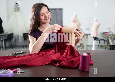 Schöne junge Dressurreiterin im Arbeitszimmer, die rotes Kleid näht Stockfoto