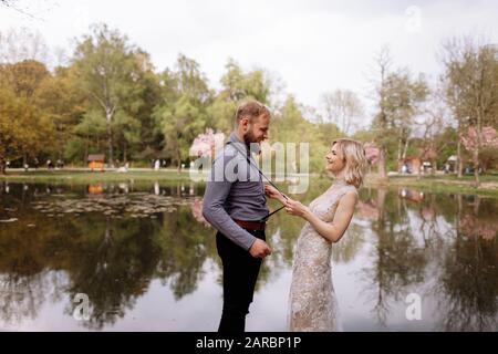 Fröhliches Hochzeitspaar hat Spaß im Frühling blühenden Sakura-Park: Frau im Luxuskleid, die bärtige zieht, bemannt Hosenträger in der Nähe des Sees. Neu eingekeilt Co Stockfoto