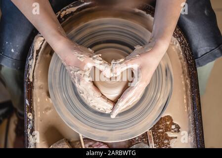 Ein Potter arbeitet in der Werkstatt mit Ton auf einem Potter-Rad. Frauenhände zeigen ein Herzschild. Das Konzept der Liebe zur Töpferkunst und Kreativität. Oben Stockfoto