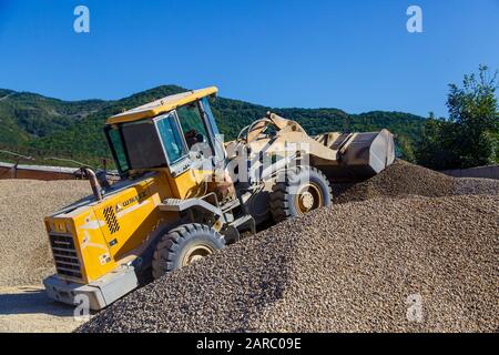 Traktorlader lädt Schutt an einem sonnigen Tag. Trümmerhaufen. Ein Traktor transportiert den zerkleinerten Stein von einem Haufen zum anderen Stockfoto