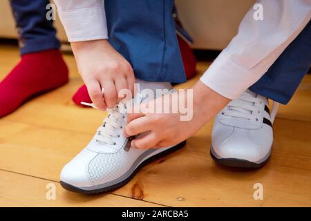 Der Junge schließt seinen weißen Schuhgarten. Nahaufnahme der Beine eines Jungen in weißen Lederschuhen auf Holzhintergrund. Schließt die Kinderschuhe. Stockfoto