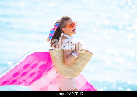 Fröhliche, moderne 40-jährige Frau in weißem T-Shirt und pinkfarbenen Shorts mit Strandabschnitt an der Küste des Ozeans, die aufblasbare Matratzen hält. Stockfoto