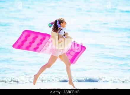 Fröhliche junge Frau in weißem T-Shirt und pinkfarbenen Shorts mit Strandabschnitt auf der Seakoast, die aufblasbare Matratze hält und läuft. Stockfoto