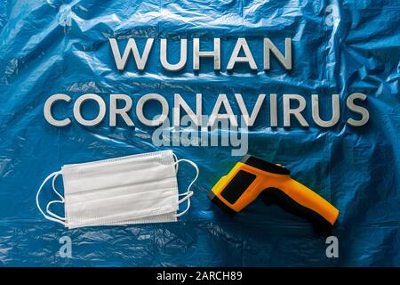 Worte wuhan Coronavirus mit Metallbuchstaben auf zerknitterter blauer Kunststoff-Filmkulisse mit Gesichtsmasken und Infrarotthermometer - flach mit Dramat Stockfoto
