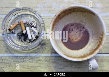 Leere Tasse Kaffee- und Zigarettenenden in Durchsichtiger Glas-Aschenbecher auf Holztischhintergrund Stockfoto