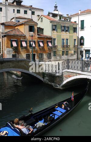 Ein Paar fährt in einer Gondel, während es unter einer Brücke an einem Kanal in Venedig, Italien, manövriert Stockfoto