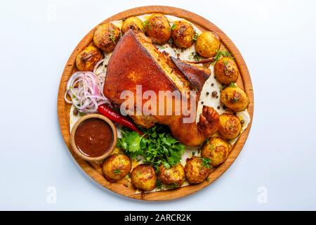 Auf einer Holzplatte liegt ein gebackener Schweinebleg, überzogen mit gebackenen Kartoffeln. Auf dem Gericht befindet sich eine Schüssel mit roter Soße, Zwiebelringen, Petersilie, Tomate. Ph Stockfoto