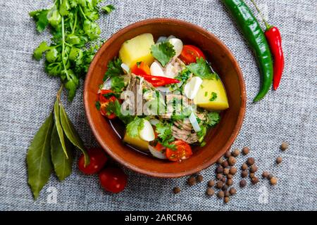Haschlama in einer Tonplatte. Zutaten: Rinderrippen, Kartoffeln, Kräuter, Knoblauch. Die Platte steht auf einer strukturierten grauen Leinwand. Um liegen Pfeffer rot und grün Stockfoto