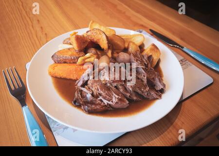 Hausgemachtes, langsam gebratenes Lamm mit Wurzelgemüse und Gravy auf einem weißen Teller auf einem Holztisch in einer Küche. Stockfoto