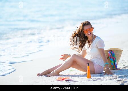 Porträt der fröhlichen jungen Frau in weißem T-Shirt und pinkfarbenen Shorts mit langen, gewellten Haaren, die am Meeresufer sitzen. Stockfoto