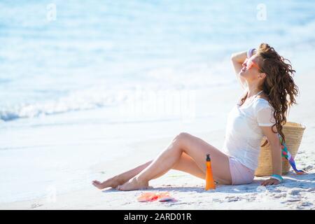 Entspannte, gesunde Frau im mittleren Alter mit weißem T-Shirt und pinkfarbenen Shorts, die auf der Seeküste sitzen. Stockfoto