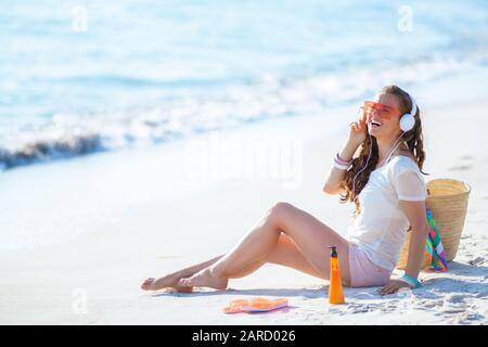Lächelnde, gesunde 40-jährige Frau in weißem T-Shirt und pinkfarbenen Shorts, die Musik mit Kopfhörern hören, während sie am Strand sitzt. Stockfoto
