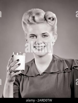 VIERZIGER JAHRE LÄCHELNDE JUNGE FRAU BLONDE HAARE UPDO MIT VICTORY ROLL LOCKEN PEARL OHRRINGE BLICK AUF DIE KAMERA HALTEN EIN GLAS MILCH - F4067 HAR001 HARS WW2 STUDIO SCHOSS GESUNDHEIT HEIM LEBEN MILCHKOPIERRAUM DAMEN PERSONEN OHRRINGE VERTRAUEN B&W AUGENKONTAKT GLÜCK KOPF UND SCHULTERN FRÖHLICHE GETRÄNKESTILE SIEG CURLS FLUID HAIRSTYLE WORLD WAR ZWEI WELTKRIEGE II VON UP SMILES UPDO VICTORY ROLLS JOYFUL STYLISH WORLD WAR 2 COIFFURE MODEN HAIRDO PROTEIN BEVERAGES BLACK AND WHITE CAUCASIAN ETHNICITY HAR001 OLD FASHIONED VICTORY ROLL Stockfoto