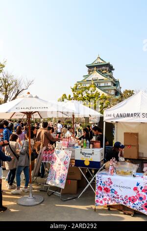 Frühlingsfest der Kirschblüte auf der Burg Osaka. Die Leute stehen Schlange, um ein Glas Chandon am Stall mit Sonnenschirmen zu kaufen. Schloss und blauer Himmel im Hintergrund Stockfoto