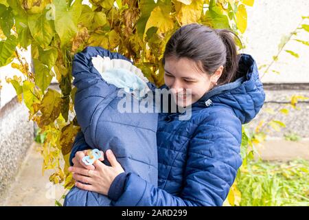 Die junge kaukasische Mutter mit einem Schnuller im Mund hält ein Neugeborenes in ihren Armen auf dem Waldhintergrund Stockfoto