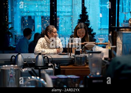 Kaukasische Und Asiatische Attraktive Frauen In Gemütlichen Pullovern, Die Lachen, Kaffee Trinken Und Im Stilvollen Café Mit Großem Fenster Sprechen. Stockfoto