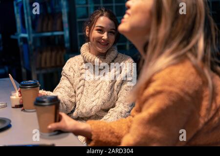 Kaukasische Und Asiatische Attraktive Frauen In Gemütlichen Pullovern, Die Beim Frühstück Kaffee Lachen Und Trinken. Konzept Der Völkerfreundschaft. Stockfoto