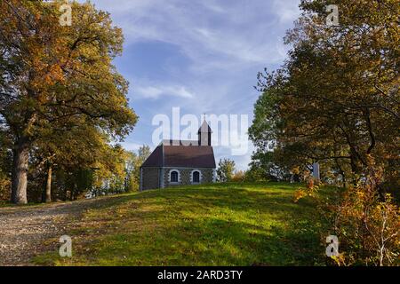 Kleine Kapelle im Wald, auf dem Hügel, mit Herbstlaub und blauem Himmel mit Wolken Stockfoto