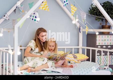 Adorable kleine Mädchen und ihre junge Mutter liest ein Buch und lächelnd während der Sitzung in eingerichteten Haus Bed Stockfoto