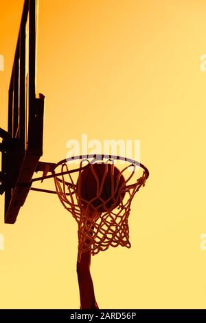 Junge Silhouetten-Basketballspieler, die hoch zum Ziel-Basketball-Hoop springen, erzielen mit einem fliegenden Slam-Dunk bei Sonnenuntergang Punkte. Erfolg und Ziel Stockfoto