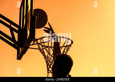 Junge Silhouetten-Basketballspieler, die hoch zum Ziel-Basketball-Hoop springen, erzielen mit einem fliegenden Slam-Dunk bei Sonnenuntergang Punkte. Erfolg und Ziel Stockfoto